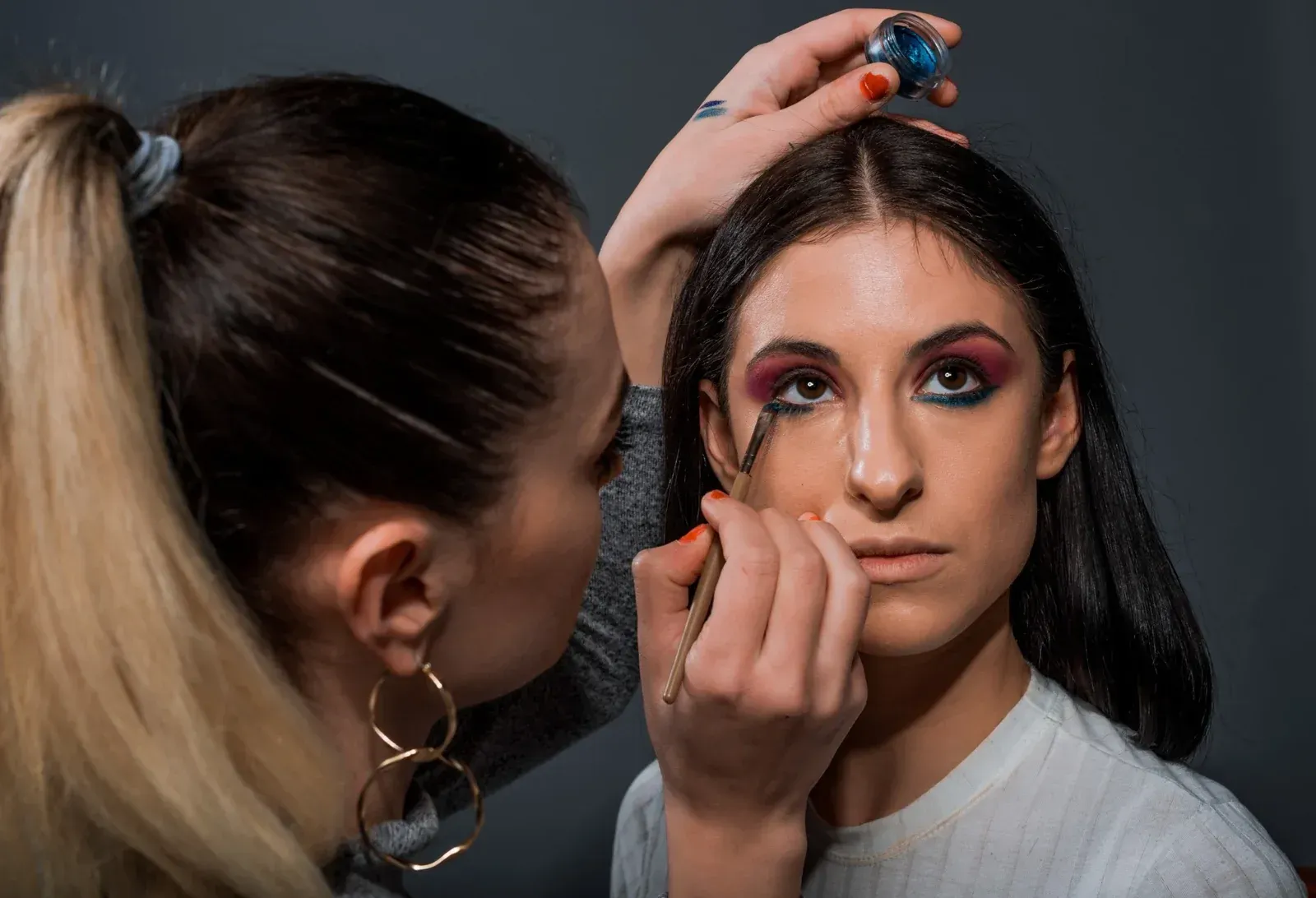 Una maquilladora aplica delineador de ojos oscuro en la línea de las pestañas inferiores de una clienta, sosteniendo un pequeño bote de maquillaje por encima de la cabeza de la clienta.