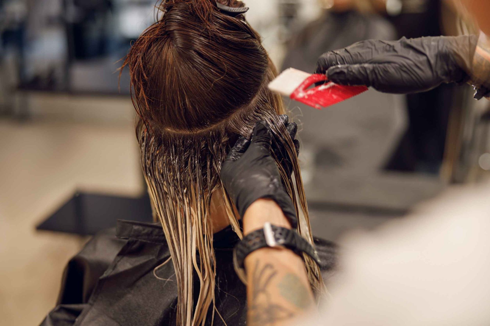 En una peluquería, una estilista con guantes negros utiliza un pincel rojo para aplicar tinte en la parte posterior de la cabeza de una persona.