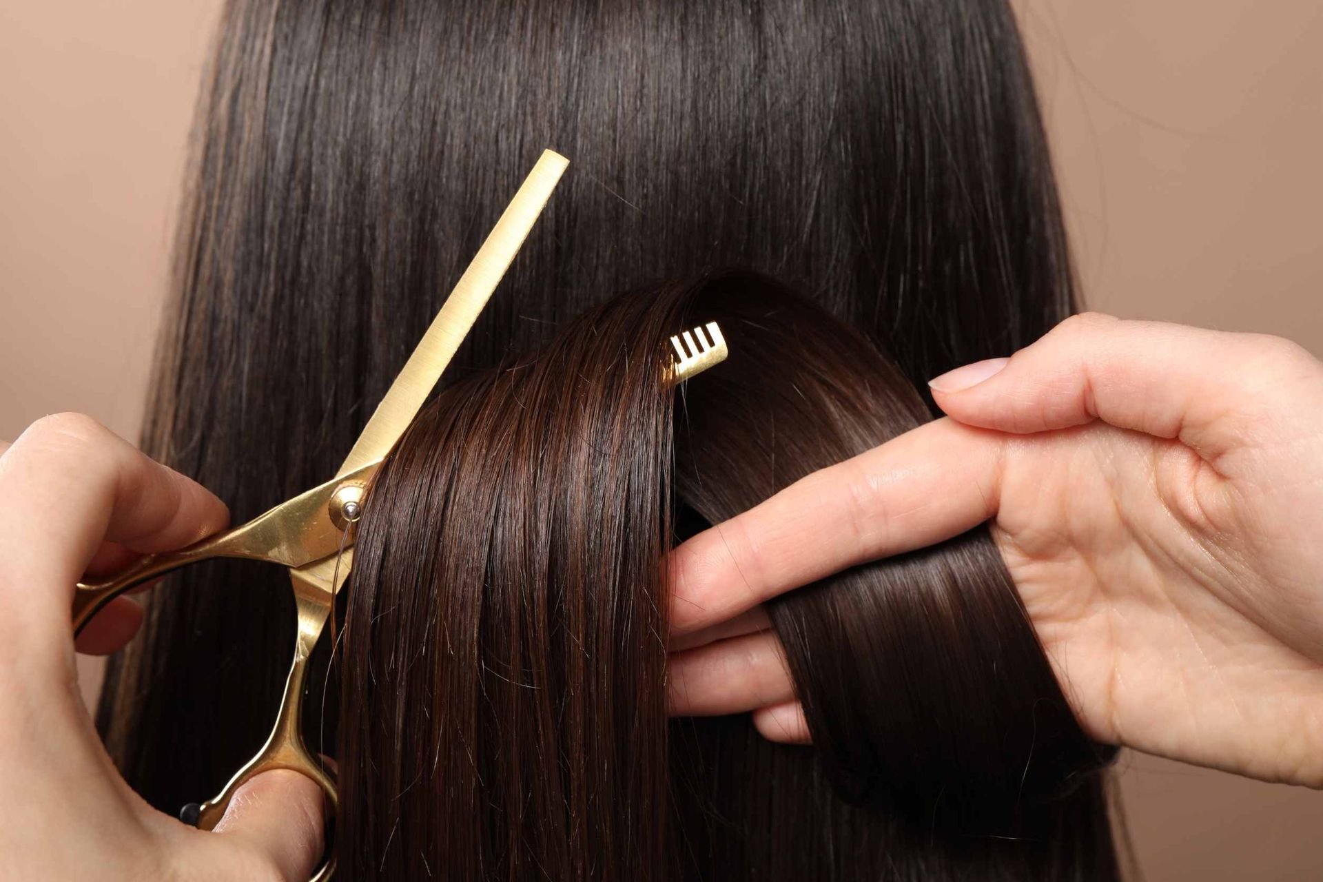 Unas manos utilizan tijeras de entresacar doradas para recortar una sección de cabello oscuro y liso.