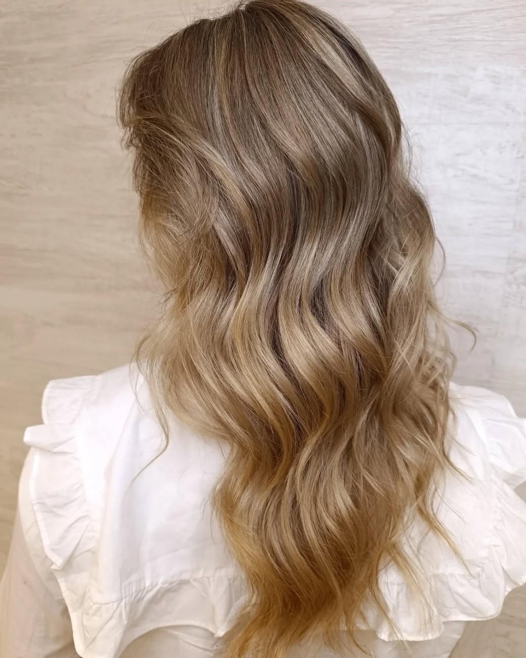 Vista posterior de una melena larga, ondulada y castaña clara con sutiles reflejos rubios sobre un fondo beige.