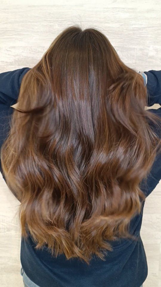 Vista posterior de una larga melena castaña ondulada con sutiles reflejos, sobre un fondo de madera clara.