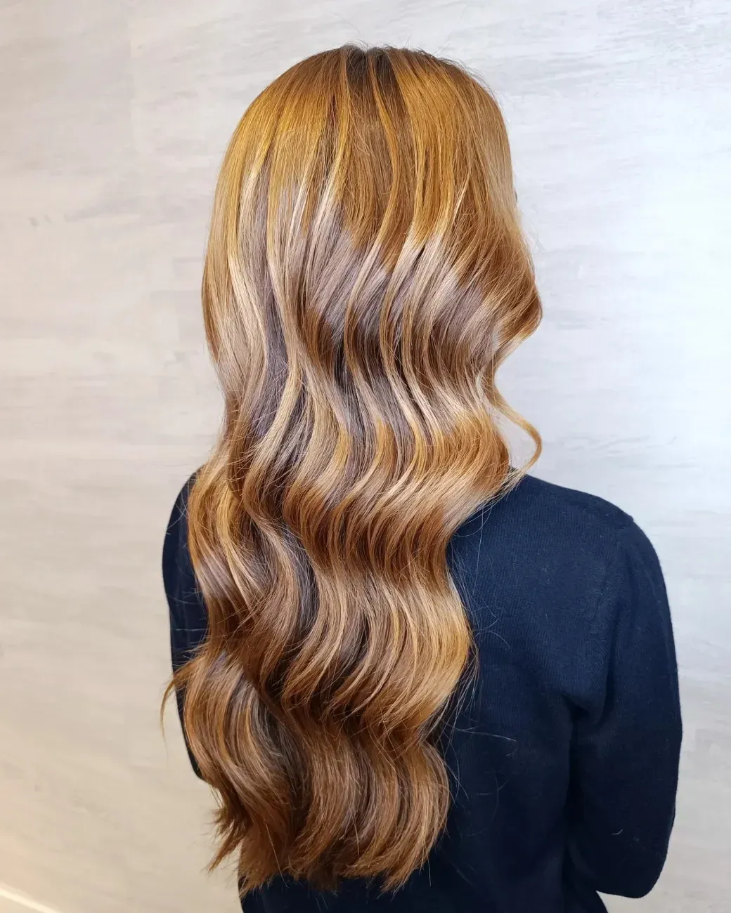 Una persona con cabello largo, ondulado y castaño caramelo, vista de espaldas contra una pared clara y lisa.