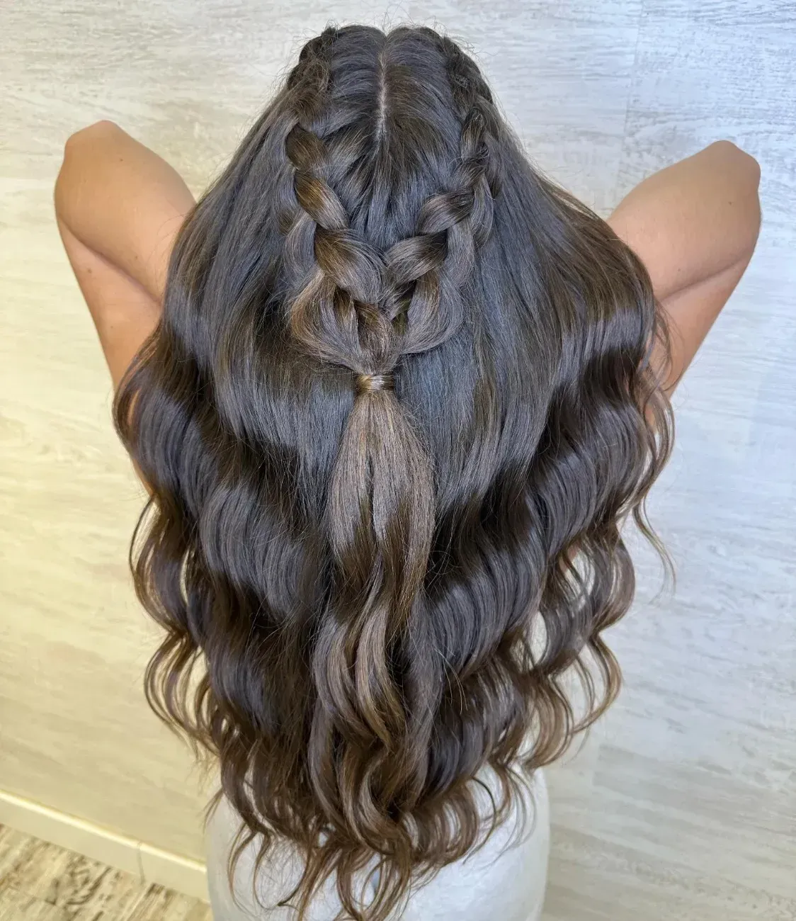 Cabello largo, ondulado y castaño, peinado con una trenza holandesa central recogida en una media coleta, contra una pared de color neutro.
