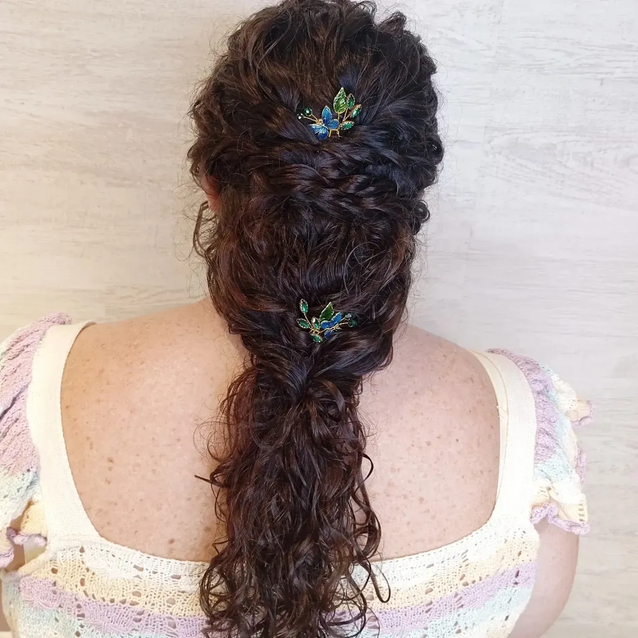 Una persona con cabello largo, ondulado y castaño oscuro, peinado en un semirecogido adornado con dos horquillas decorativas de flores azules y verdes.