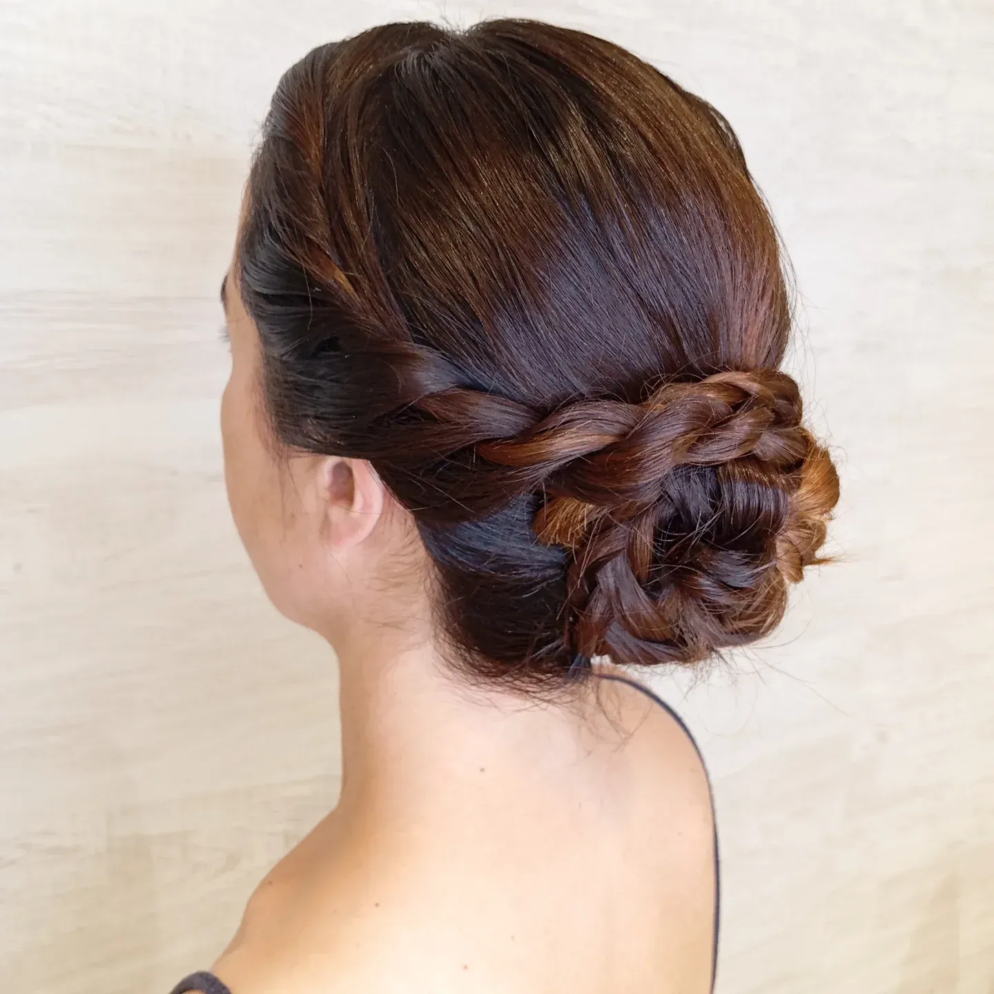 Una persona de cabello castaño oscuro con un elaborado peinado de moño trenzado, vista de espaldas contra un fondo de madera clara.