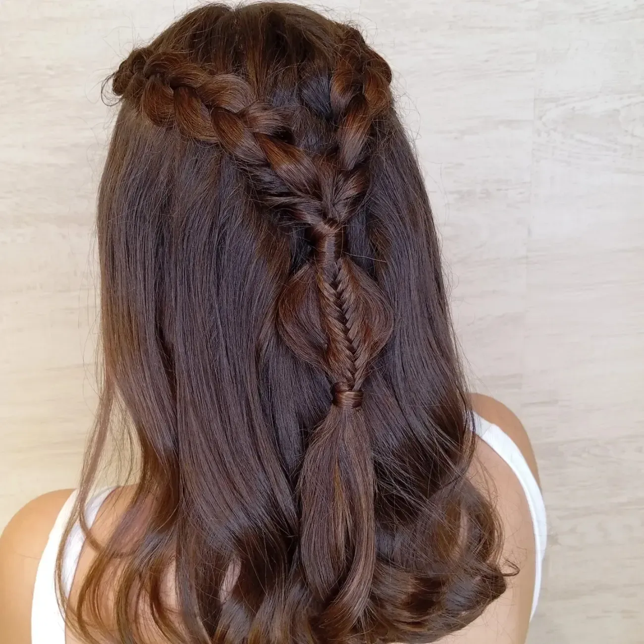 Una persona con cabello largo, oscuro y ondulado, peinado en un semirecogido con una corona trenzada y una trenza decorativa que se introduce a través del cabello.