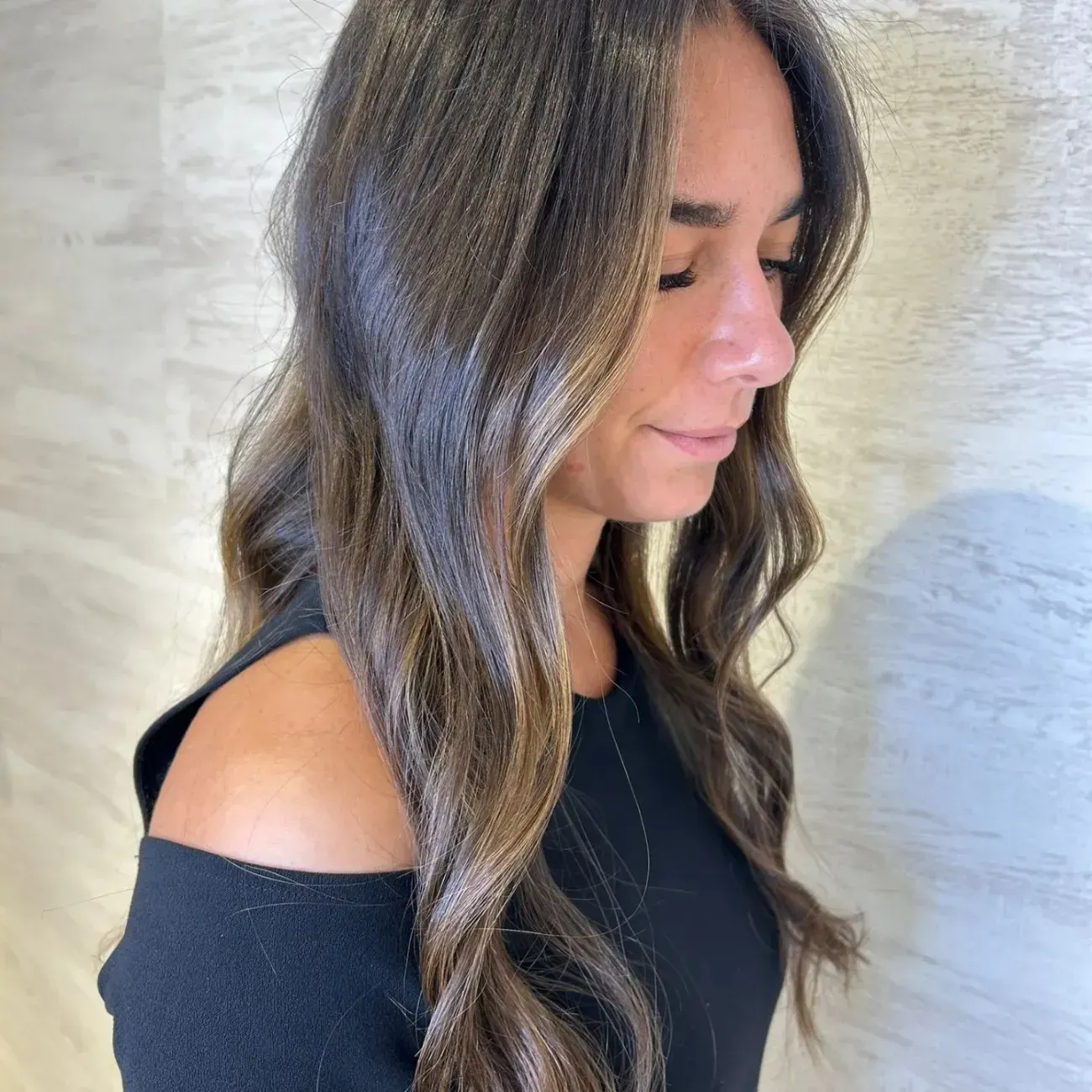 Una persona con una blusa negra, cabello largo, ondulado y castaño oscuro con reflejos más claros, vista de perfil contra una pared clara.