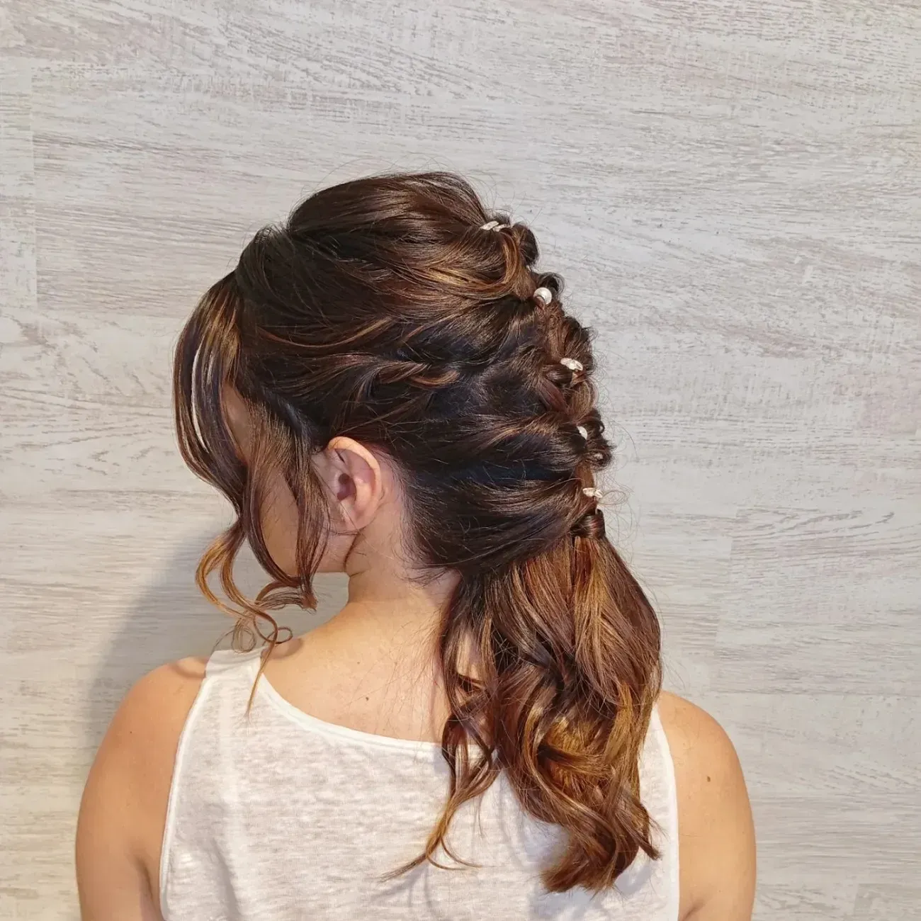 Cabello castaño recogido en una trenza texturizada a media altura, adornada con pequeñas horquillas decorativas, visto desde atrás contra una pared de madera.