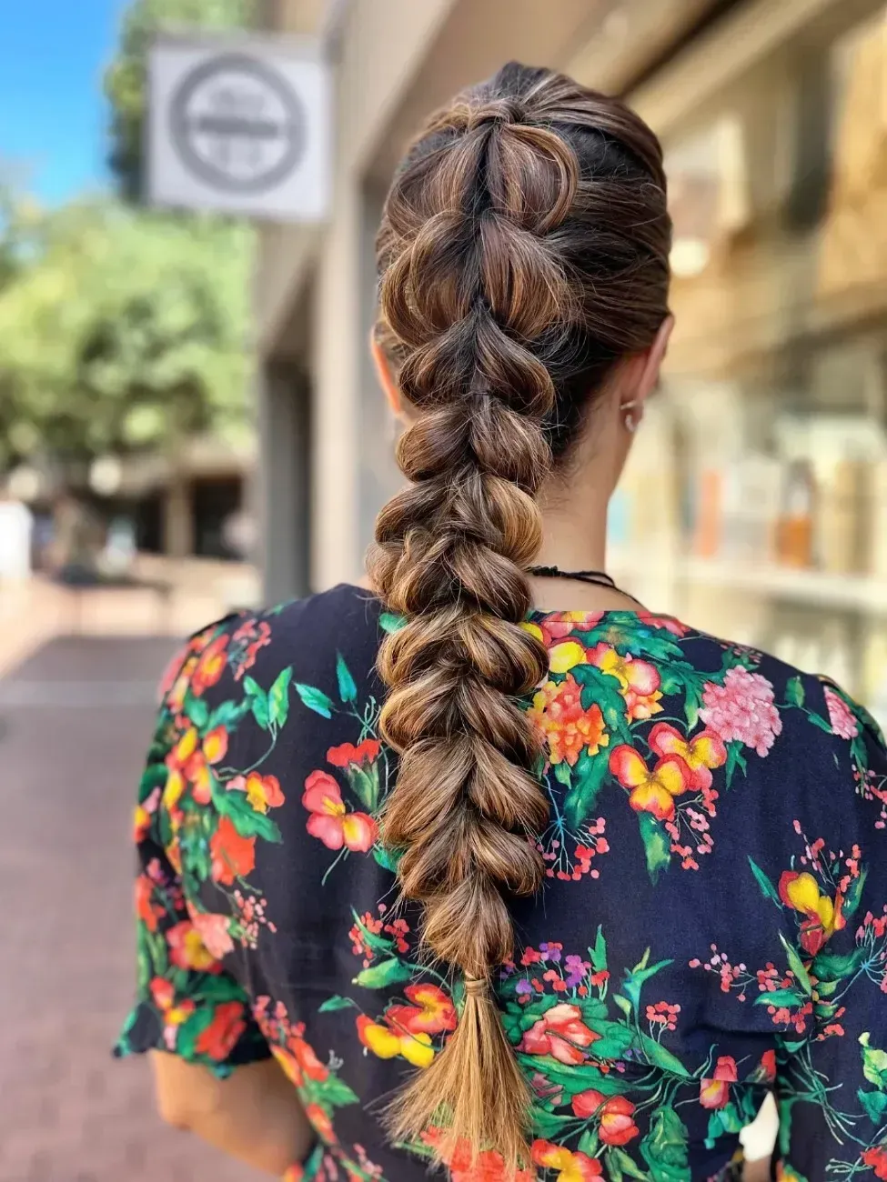 Una persona con el pelo castaño con mechas recogido en una trenza gruesa y voluminosa, vestida con un vestido floral, caminando por la acera.