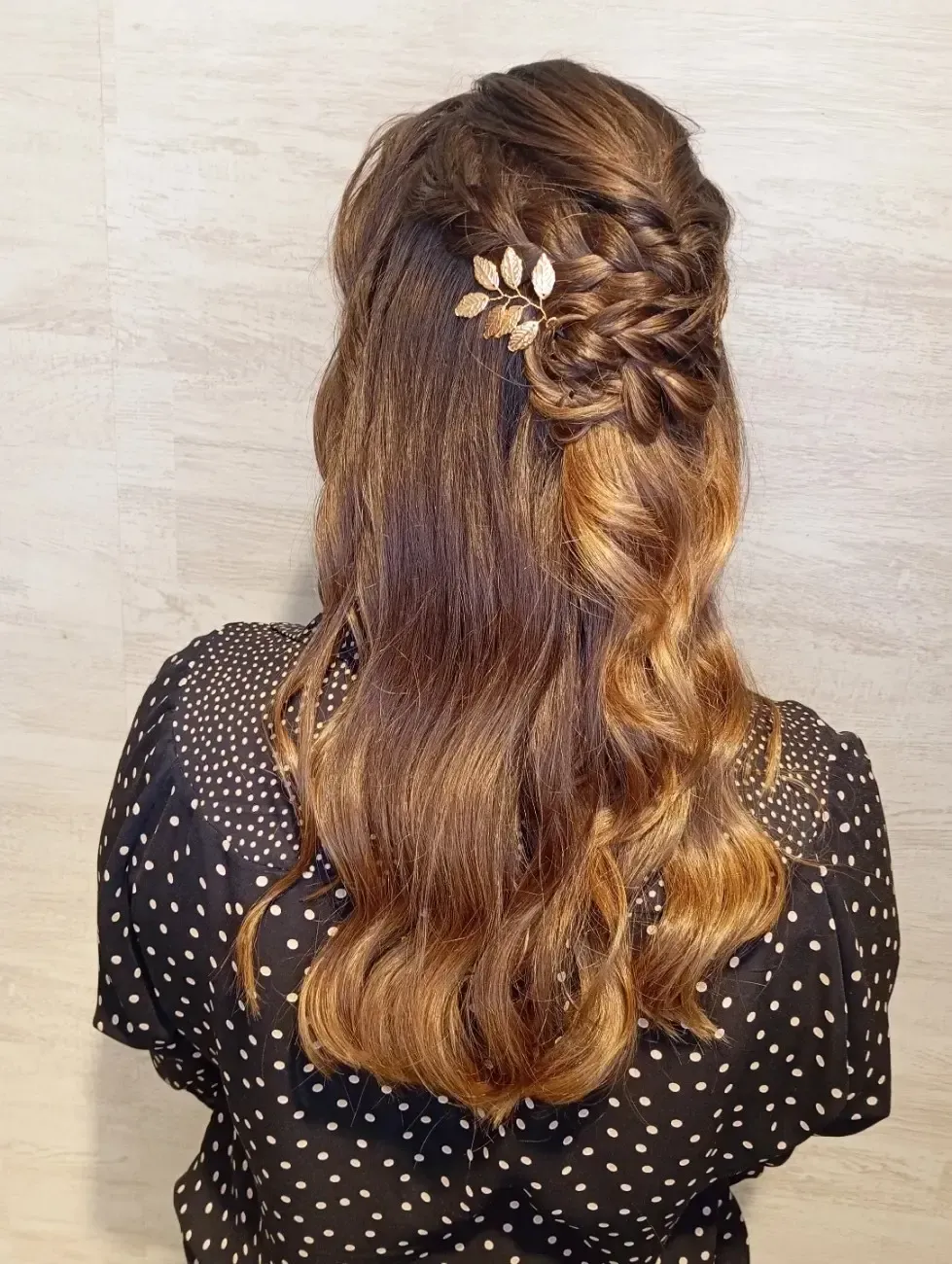 Una persona con el cabello ondulado y con mechas, peinado en un semirecogido trenzado y sujeto con un accesorio para el cabello dorado en forma de hoja.