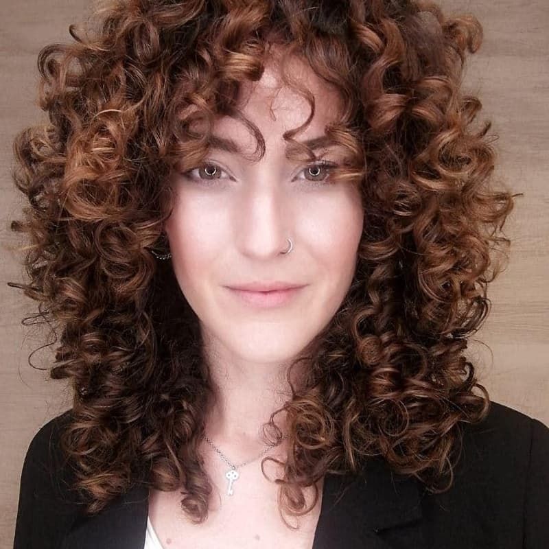 Una persona con cabello castaño largo, abundante y rizado, con flequillo, que viste un blazer negro y un collar delicado.