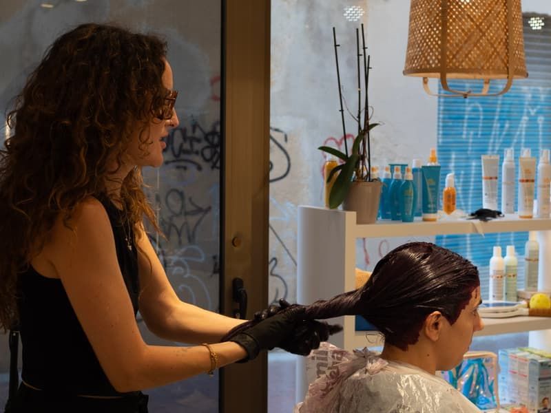 Una estilista con guantes negros aplica tinte al cabello de una clienta en un salón de belleza, con estantes de productos para el cabello al fondo.