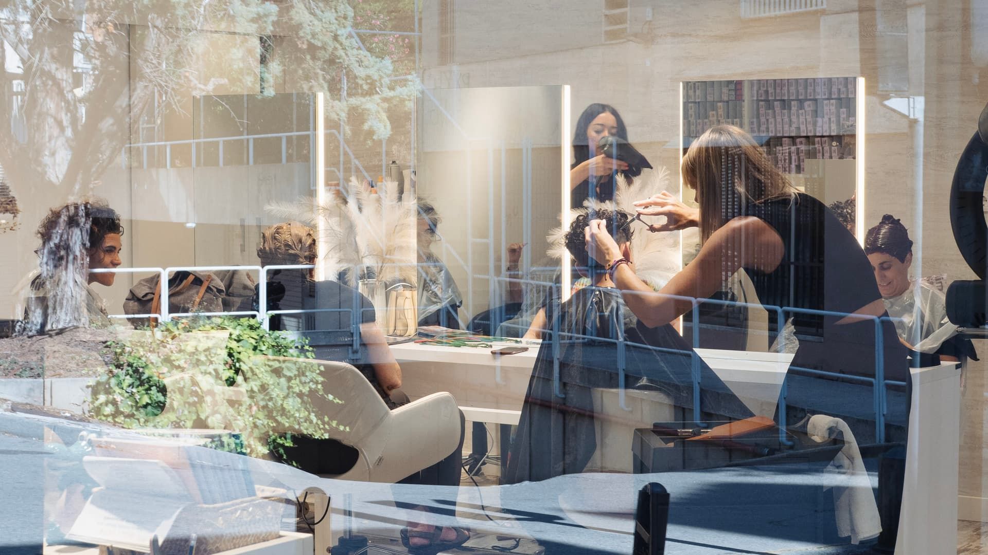 Una estilista corta el cabello de una clienta en un salón con múltiples espejos verticales que reflejan el interior y los árboles del exterior.