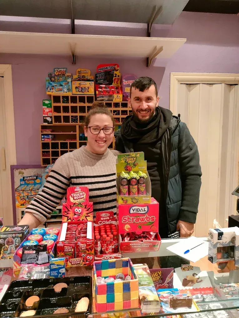 Dos personas sonriendo detrás de un mostrador de dulces lleno de golosinas coloridas.