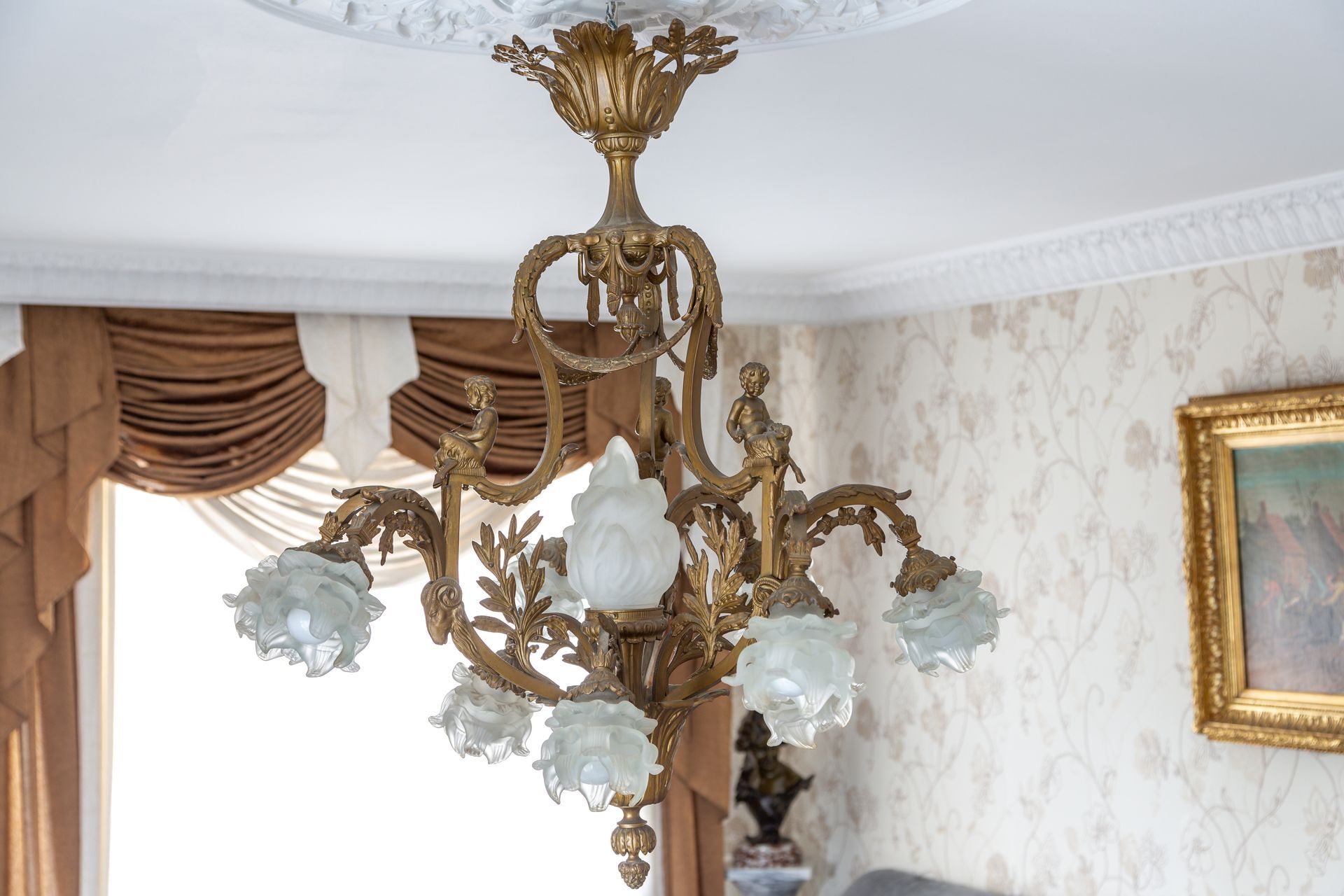 Un lustre doré orné de six abat-jour en verre dépoli est suspendu à une rosace de plafond blanche finement travaillée