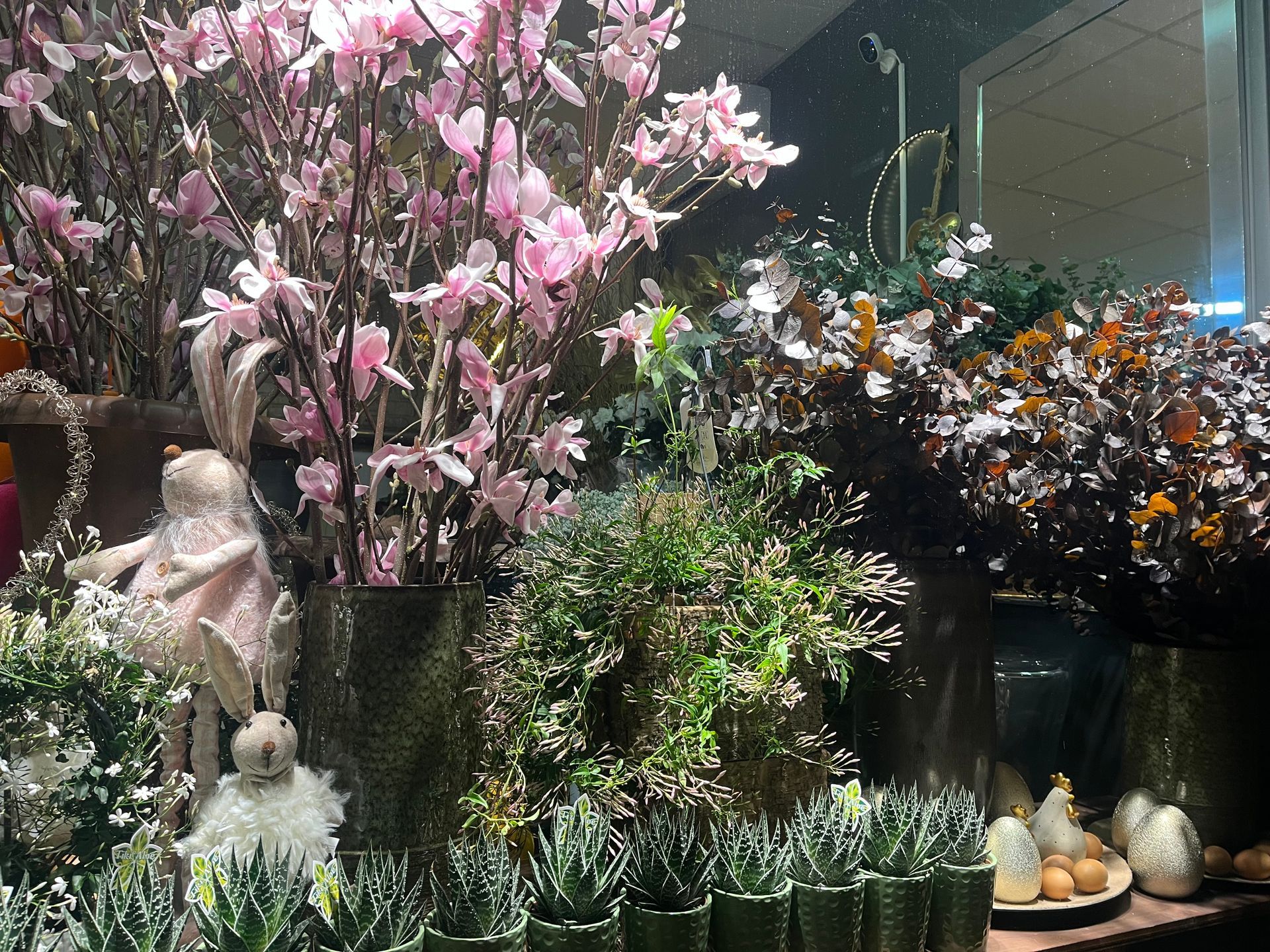 Fleurs roses et plantes en pot exposées dans une jardinerie, avec des statues et des fleurs décoratives.