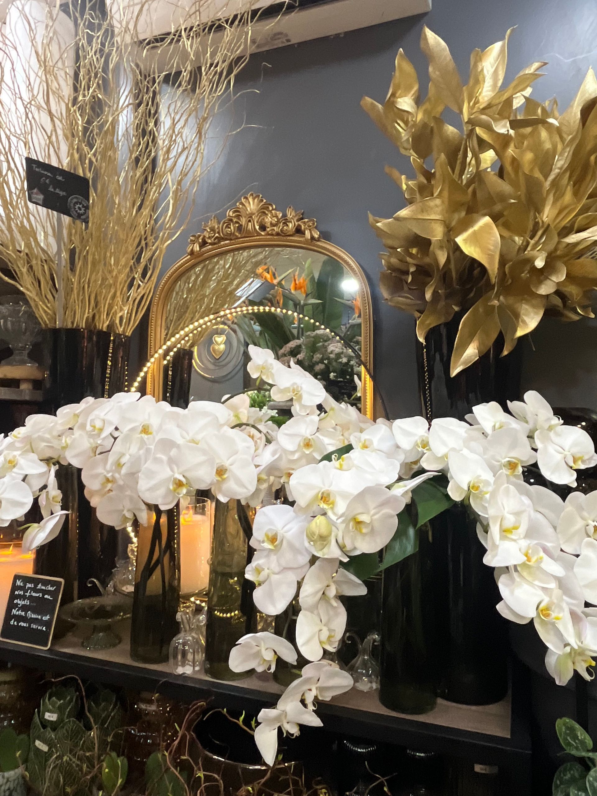 Des orchidées blanches et des compositions florales dorées sont disposées sur une table à côté d'un miroir doré orné, dans une vitrine.