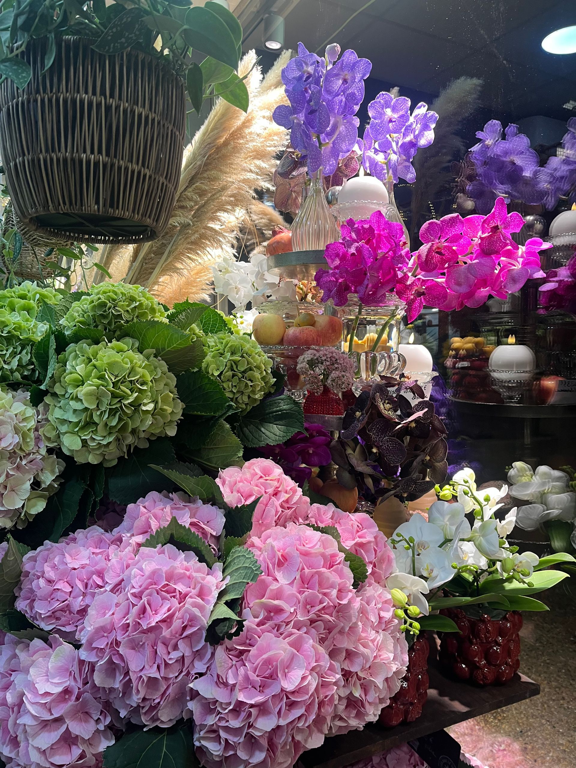 Présentation de fleurs colorées avec des fleurs roses, violettes, vertes et blanches dans un marché ou une boutique couverte.