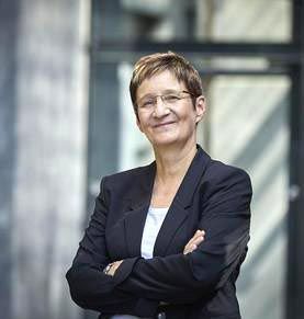 Eine Frau in einem schwarzen Anzug und mit Brille steht mit verschränkten Armen da und lächelt.