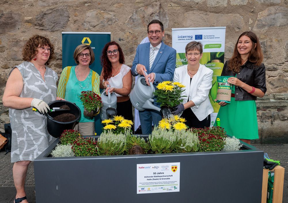 Five adults pose with potted flowers and a planter box in front of a stone wall.