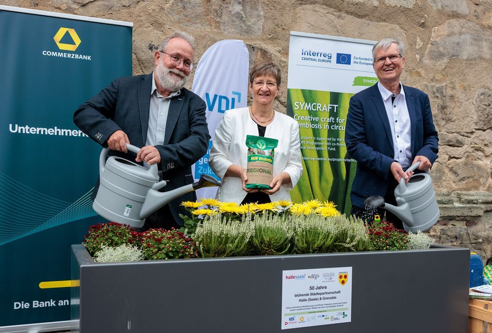 Three people smiling with gardening tools and a potted plant at an outdoor event display