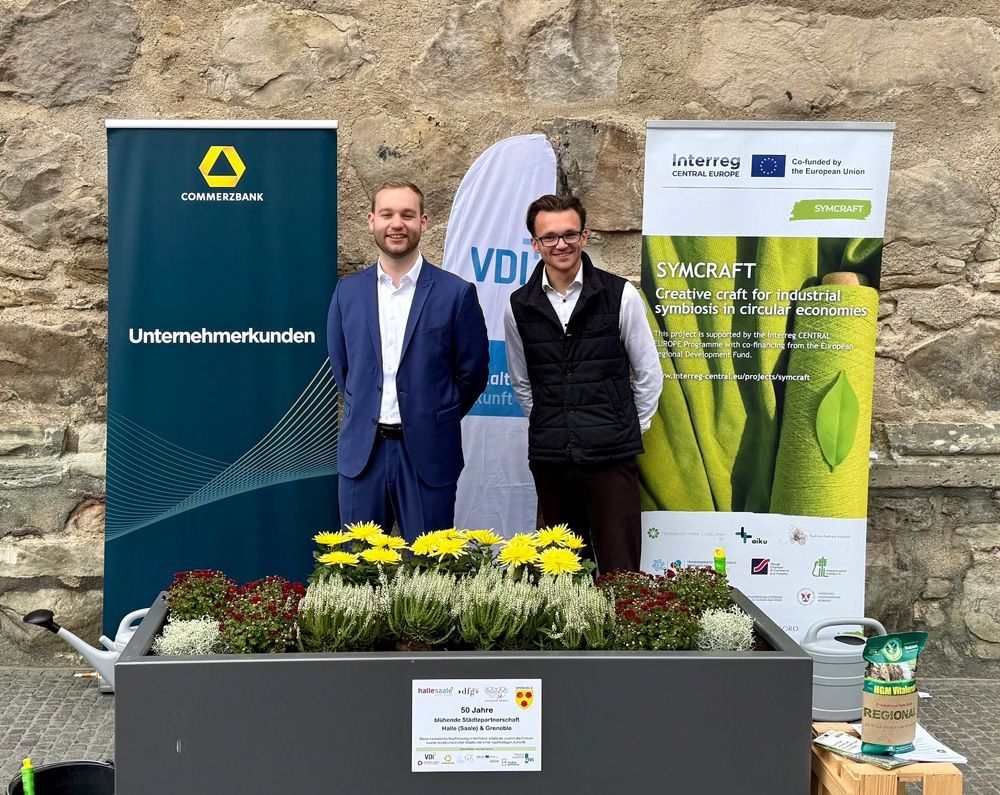 Two men stand behind a flower planter at an outdoor trade booth with green promotional banners.