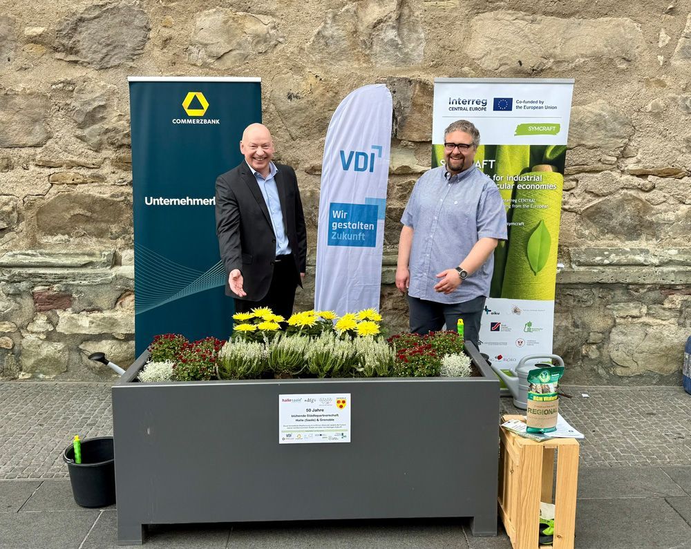 Two men standing beside a flower planter and sponsor banners against a stone wall outdoors