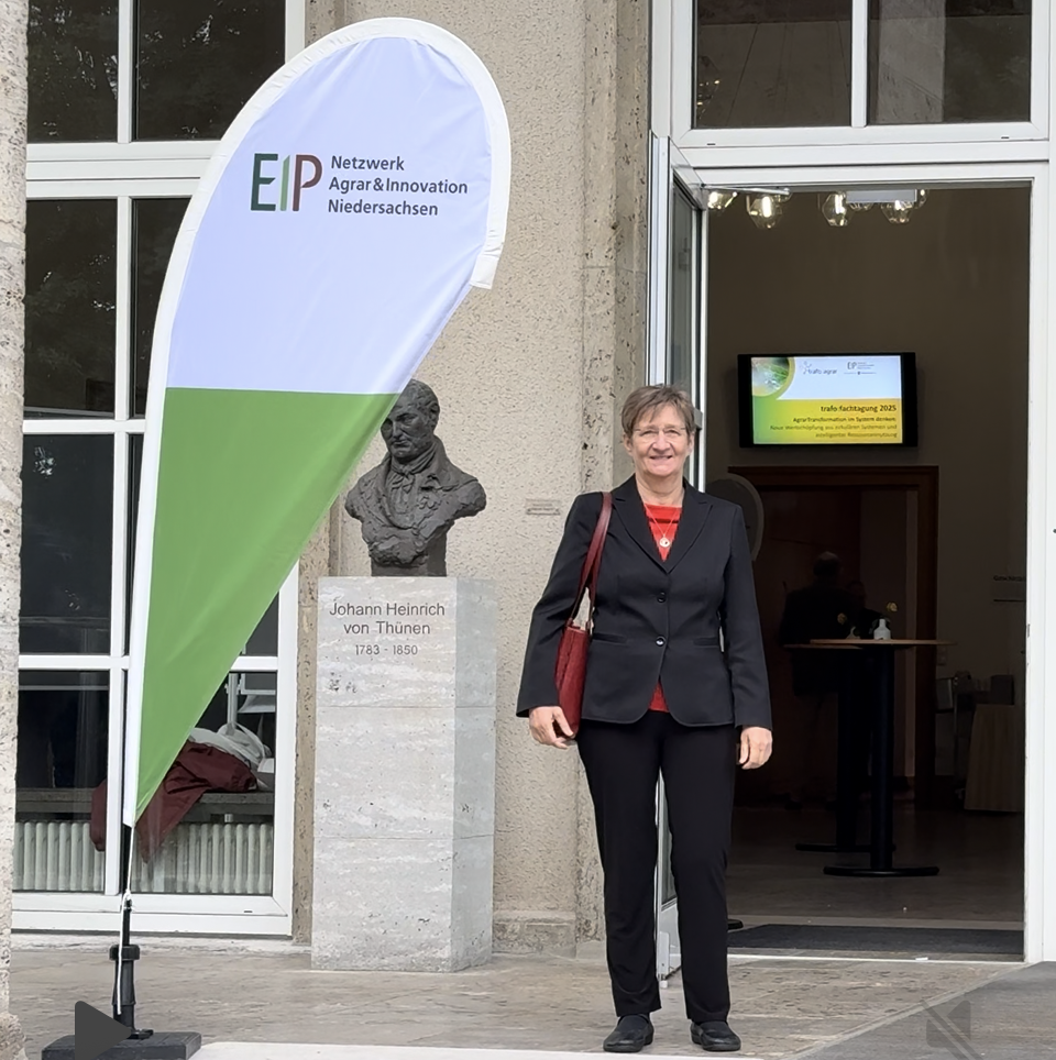 Frau im schwarzen Anzug steht mit einer Veranstaltungsflagge und einer Büste vor einem Gebäudeeingang.