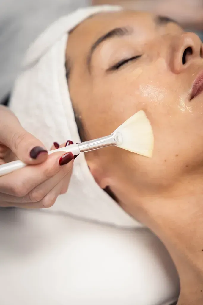 Tratamiento facial con un pincel aplicando un producto en la mejilla de una mujer mientras está tumbada en una camilla de spa