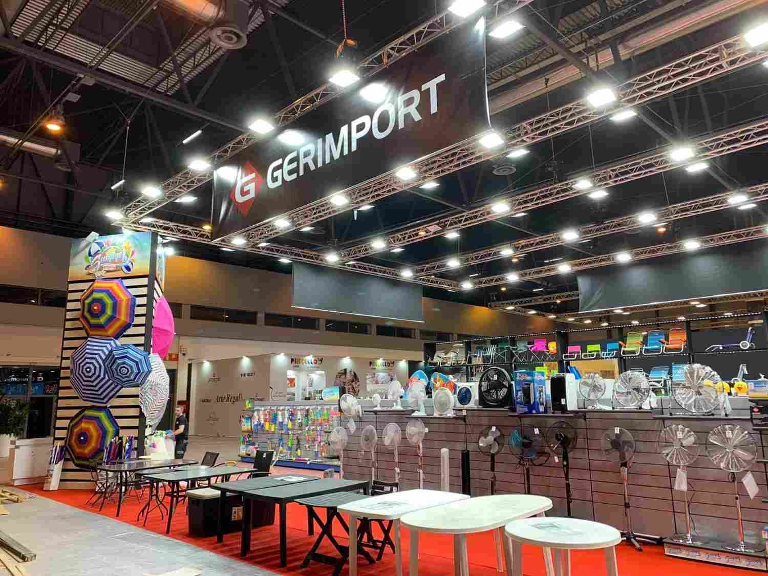 Stand de GERIMPORT en una feria, con luces colgantes, expositores apilados y coloridos, y mesas con productos