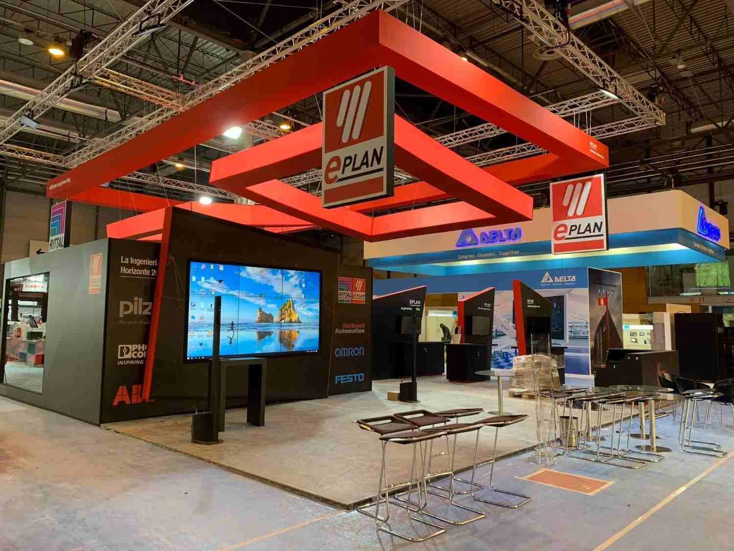 Stand de feria con una estructura superior roja, grandes pantallas digitales y una zona de asientos en el recinto ferial