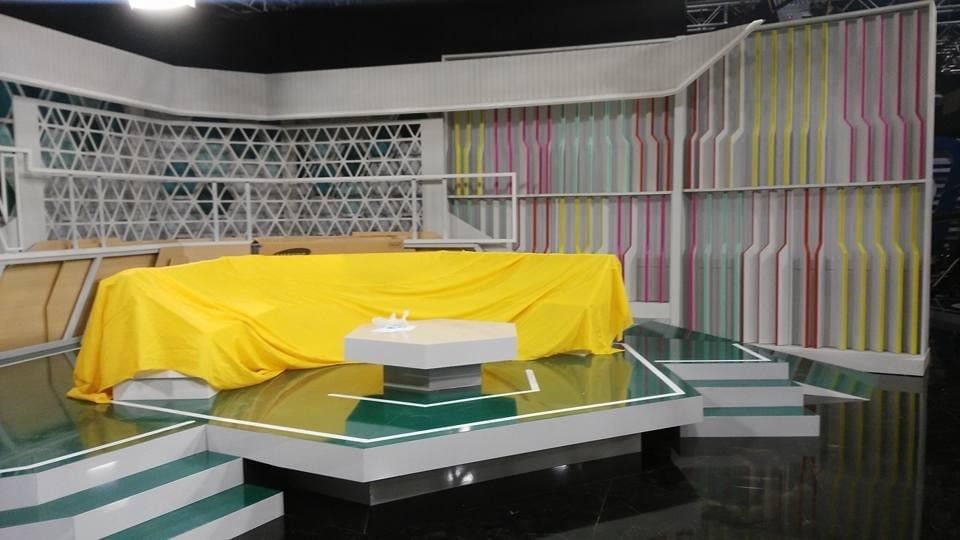 Plató de televisión con una mesa cubierta con un mantel amarillo, una silla blanca y un fondo de rayas en tonos pastel