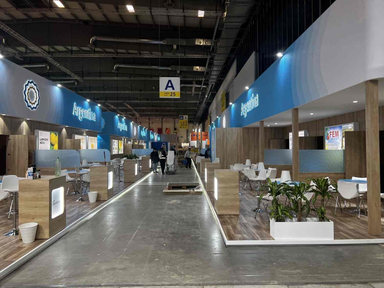 Pabellón de exposiciones interior con puestos azules, zonas de descanso y un amplio pasillo central bien iluminado