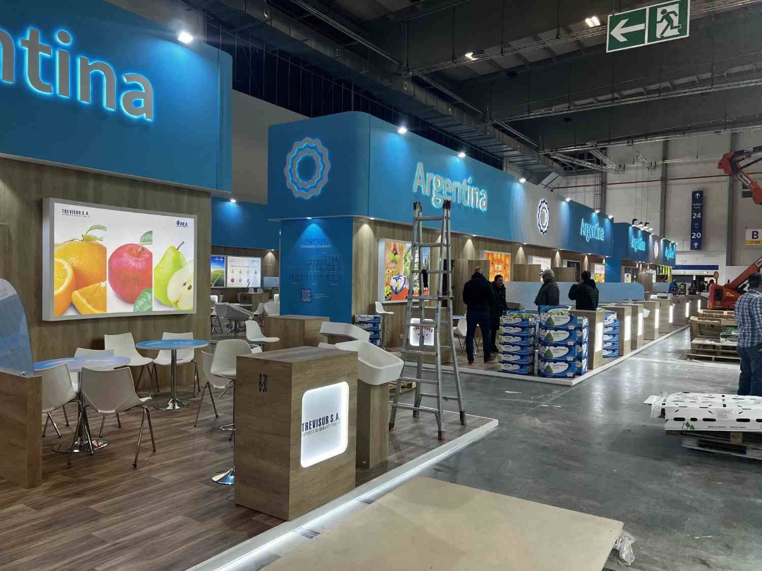 Pabellón de exposiciones interior con puestos azules, asientos y mesas de exhibición a lo largo de un pasillo