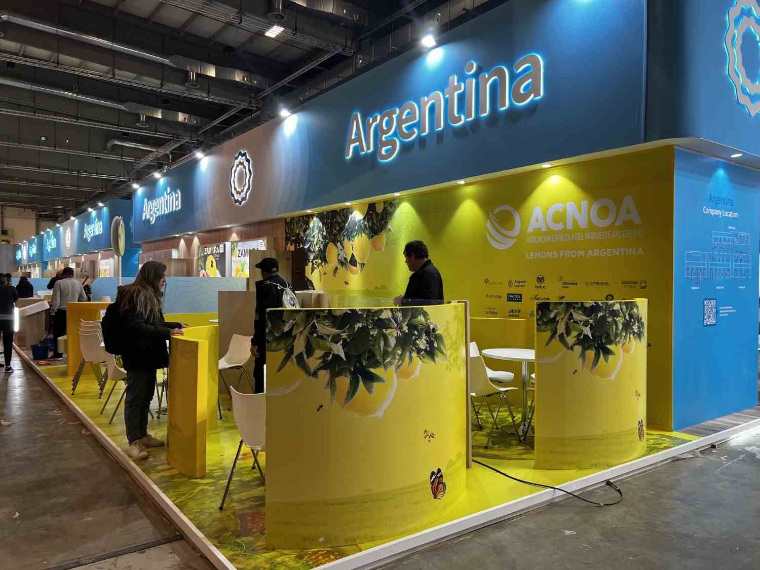 Stand de Argentina con paredes azules y amarillas, personas de pie frente a mostradores en un centro de convenciones