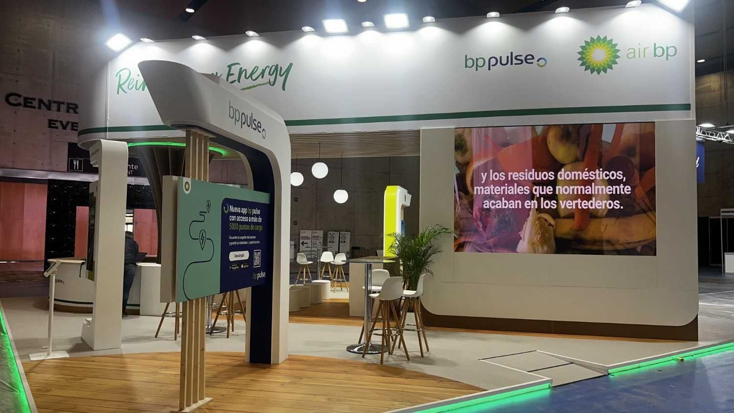 Stand de feria comercial con imagen de marca en blanco y verde, paneles curvos y una pantalla promocional iluminada.