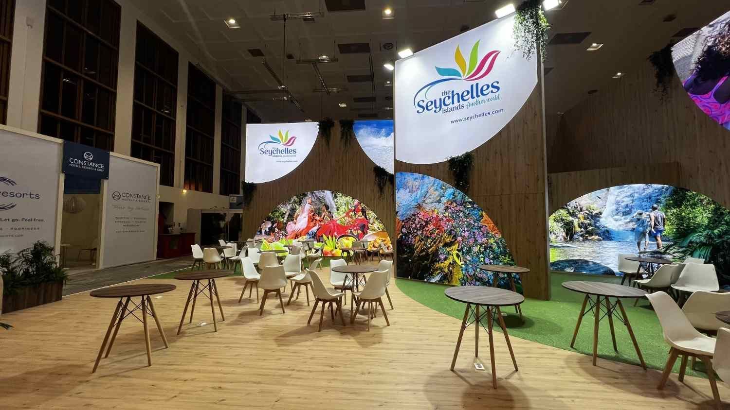Stand para eventos en interiores con adornos de hojas de colores, mesas, sillas y letreros colgantes en un amplio salón