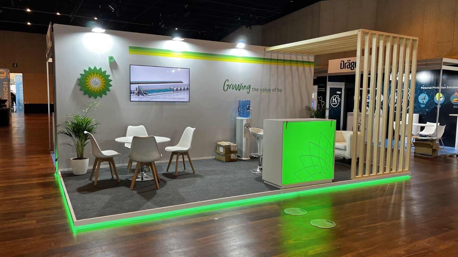 Stand moderno para feria comercial, con sillones blancos, iluminación verde de acento y paredes de listones de madera