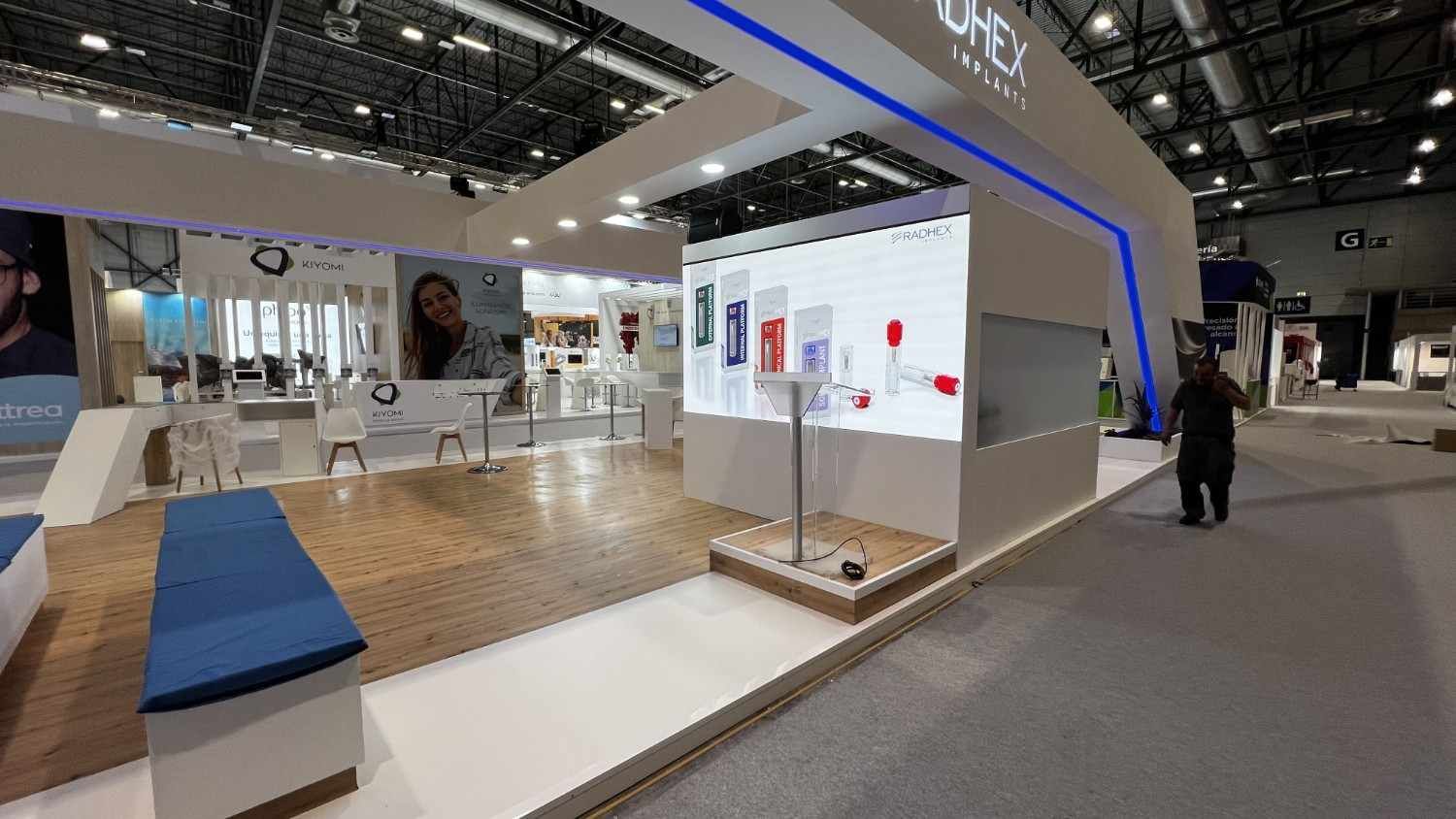 Moderno stand de feria comercial con expositores blancos, un arco iluminado en azul y gente pasando por el recinto ferial