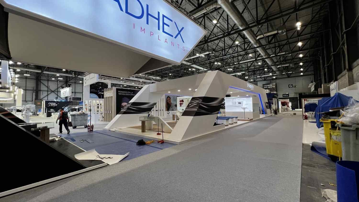 Stand de feria vacío con un expositor geométrico blanco y una pancarta de ADHEX en un gran pabellón de convenciones