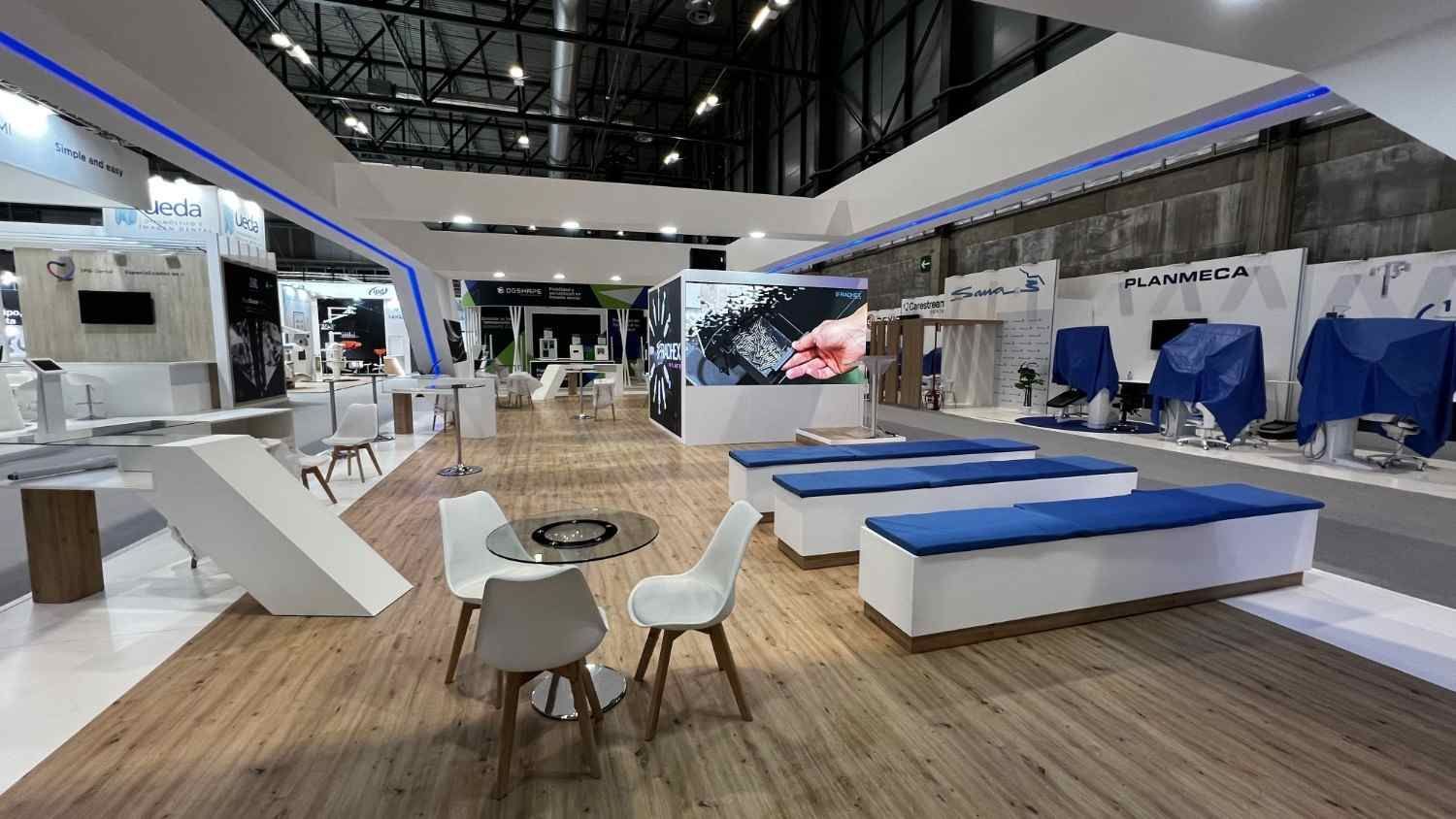 Stand de exposición moderno con expositores blancos, detalles azules, zona de descanso y suelo de madera en un gran pabellón