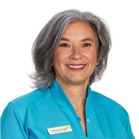 Mulher de cabelos grisalhos sorri, vestindo uma blusa turquesa com uma etiqueta de nome.