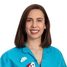 Mulher de cabelos castanhos, sorrindo, vestindo uma camisa azul com detalhes em forma de nuvem e dentes.