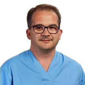 Homem de óculos e uniforme cirúrgico azul, olhando para a câmera.