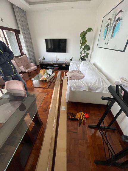Sala de estar iluminada com sofá branco, TV, plantas, piso de madeira e uma pessoa perto da janela.