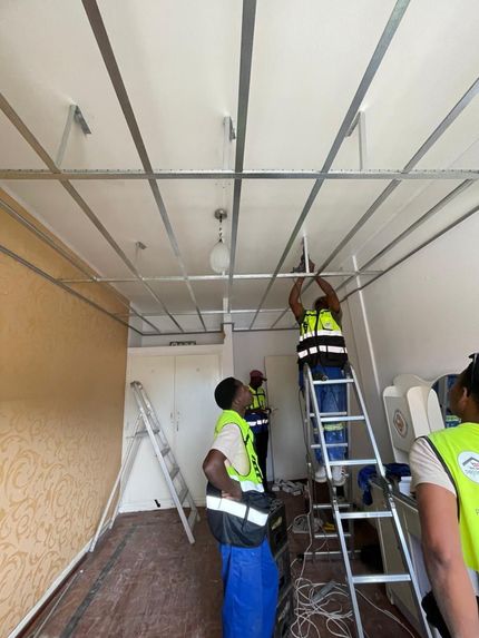 Operários instalando uma estrutura de teto dentro de uma sala, um deles em uma escada enquanto outros o auxiliam.