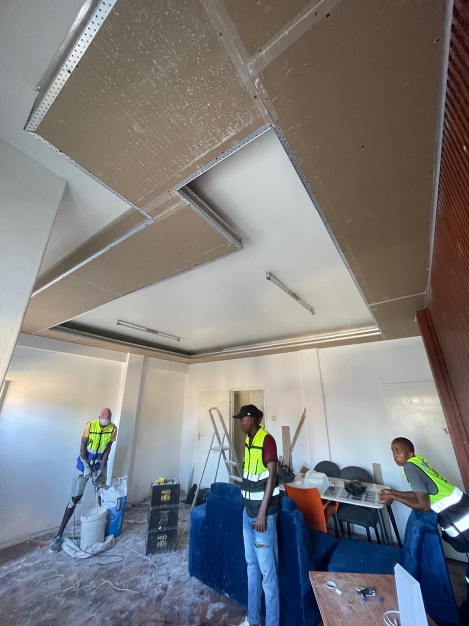 Operários instalando drywall e painéis de teto em uma sala em construção, com ferramentas e materiais ao redor.