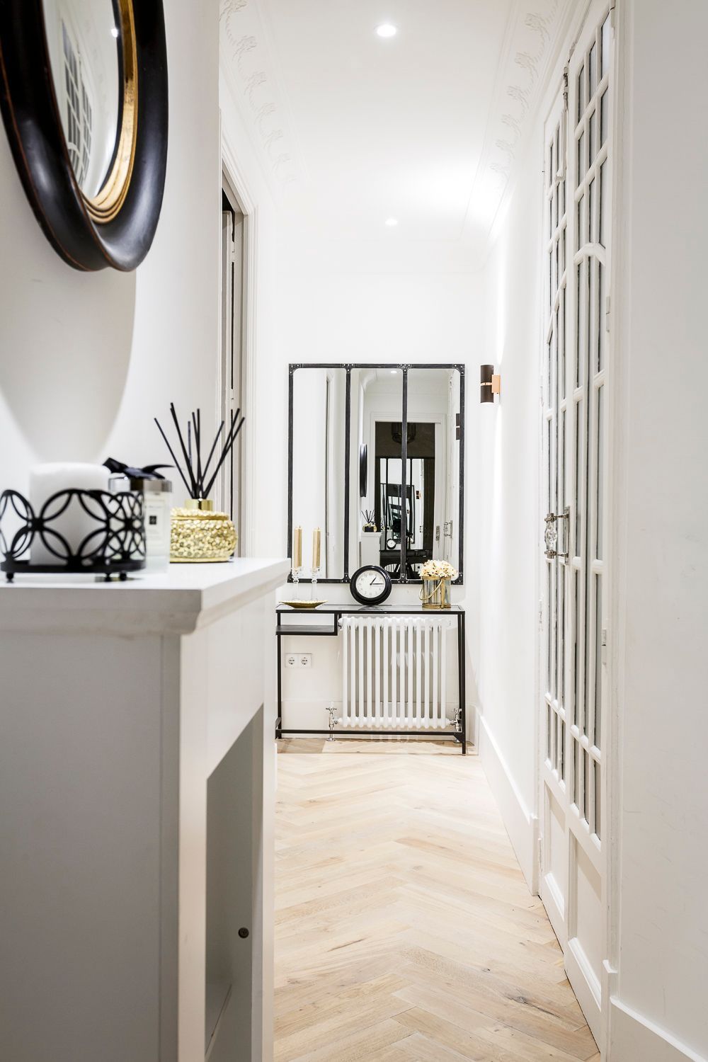 Pasillo estrecho y blanco con suelo de madera en espiga, mesa consola, espejo y elementos decorativos.