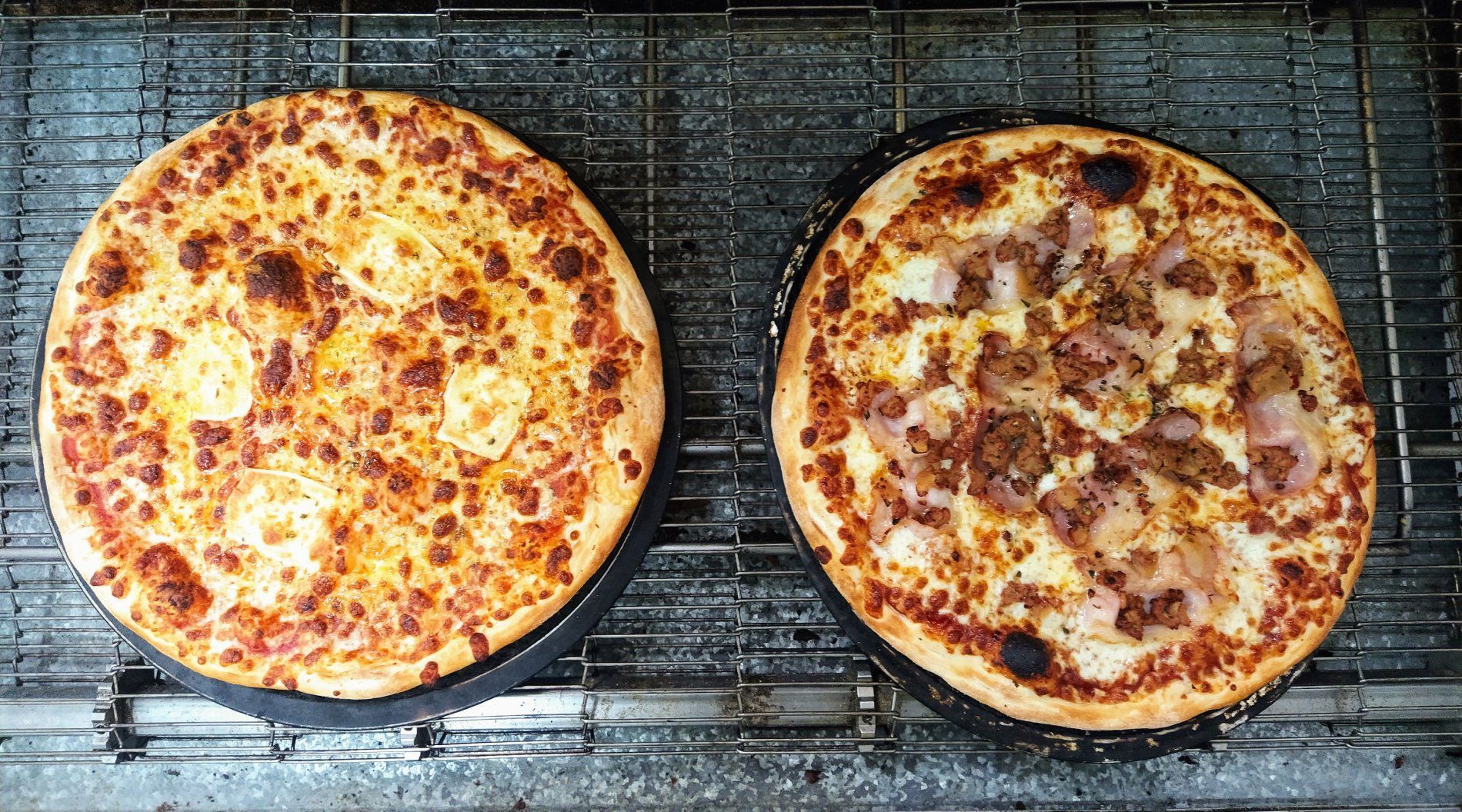Dos pizzas sobre piedras negras sobre una rejilla metálica. Una lleva queso, la otra carne y cebolla.