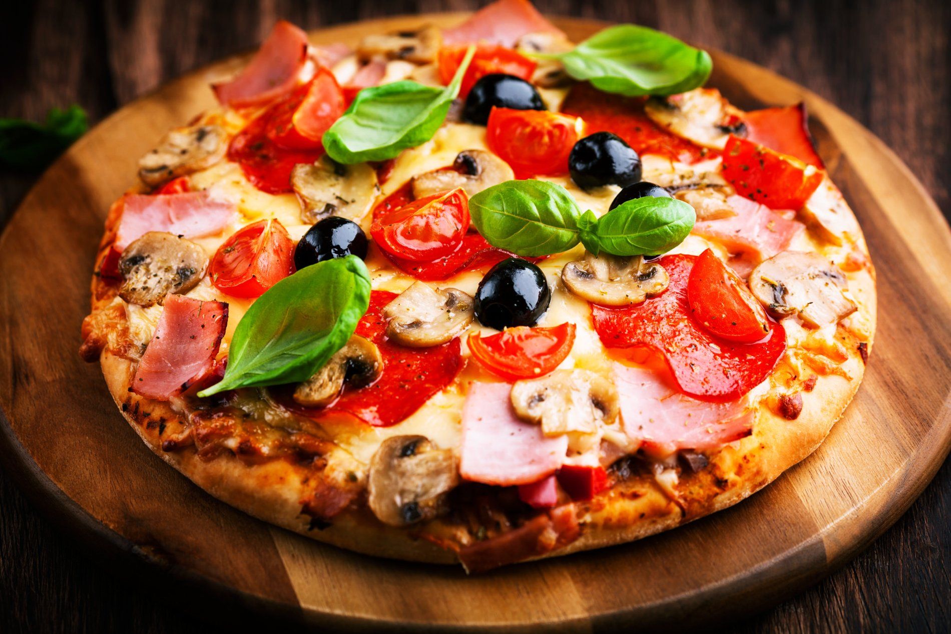 Pizza sobre tabla de madera, cubierta con tomates, aceitunas, champiñones, jamón y albahaca.